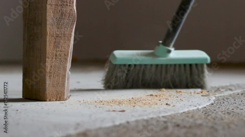 A broom sweeping a dusty floor in an unfinished room. A table leg and visible dust motion create a feeling of active cleaning in progress.