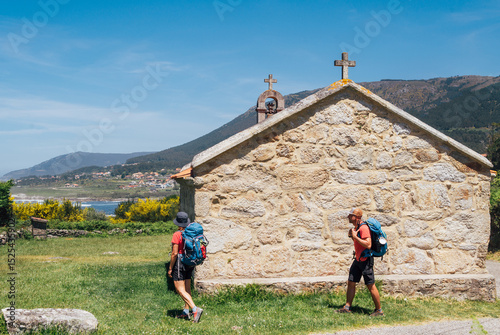 Fototapet Couple of pilgrims with backpacks walking next to old church on Camino Portuguese Way, inspiring walk on famous Camino de Santiago