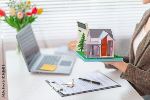 Inside a real estate office a real estate agent is using a house mockup to present the design and promotions that the client will receive when they sign sales contract. Real estate investment concept