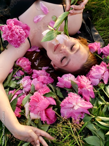 Happy smiling young woman in summer outdoors in a field with pink peony flowers enjoying life in a moment at sunset