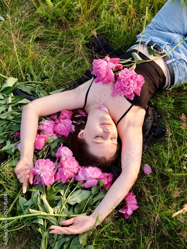 Happy smiling young woman in summer outdoors in a field with pink peony flowers enjoying life in a moment at sunset