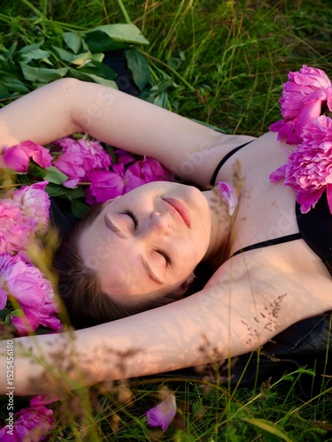 Happy smiling young woman in summer outdoors in a field with pink peony flowers enjoying life in a moment at sunset