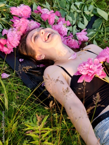 Happy smiling young woman in summer outdoors in a field with pink peony flowers enjoying life in a moment at sunset