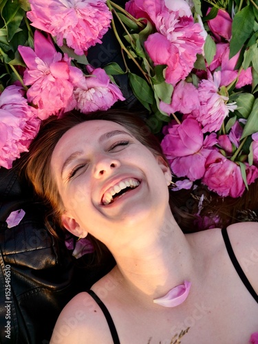 Happy smiling young woman in summer outdoors in a field with pink peony flowers enjoying life in a moment at sunset