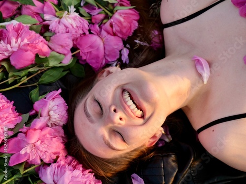 Happy smiling young woman in summer outdoors in a field with pink peony flowers enjoying life in a moment at sunset