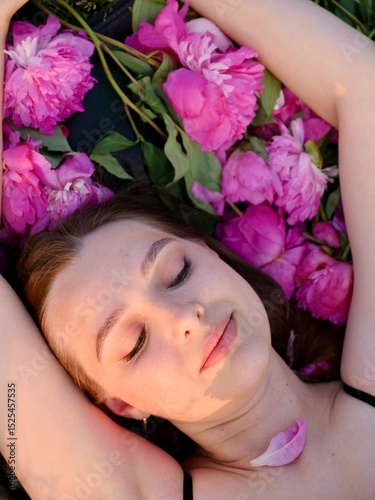 Happy smiling young woman in summer outdoors in a field with pink peony flowers enjoying life in a moment at sunset