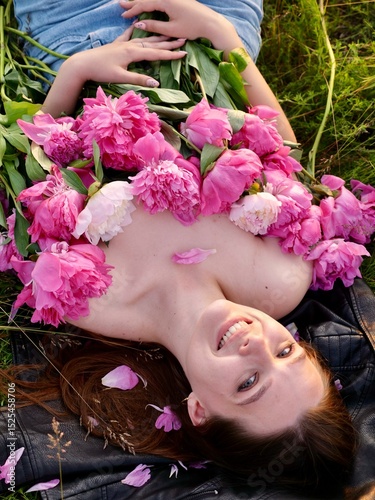 Happy smiling young woman in summer outdoors in a field with pink peony flowers enjoying life in a moment at sunset