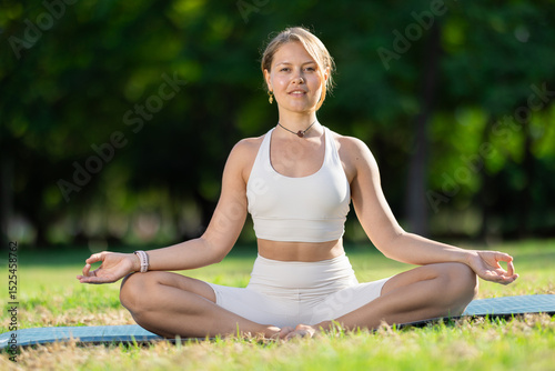 Girl fulfill lotus position, padmasana during yoga workout in city park, on green lawn. Concept of balance, relaxation, physical health, longevity