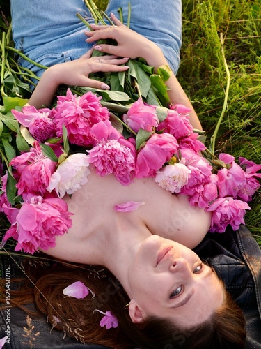 Happy smiling young woman in summer outdoors in a field with pink peony flowers enjoying life in a moment at sunset