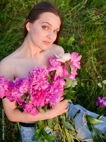 Happy smiling young woman in summer outdoors in a field with pink peony flowers enjoying life in a moment at sunset