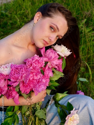 Happy smiling young woman in summer outdoors in a field with pink peony flowers enjoying life in a moment at sunset