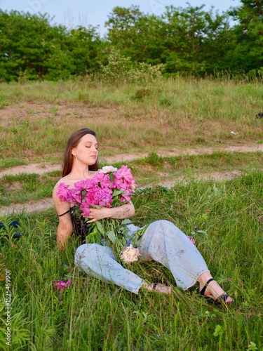 Happy smiling young woman in summer outdoors in a field with pink peony flowers enjoying life in a moment at sunset