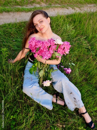 Happy smiling young woman in summer outdoors in a field with pink peony flowers enjoying life in a moment at sunset