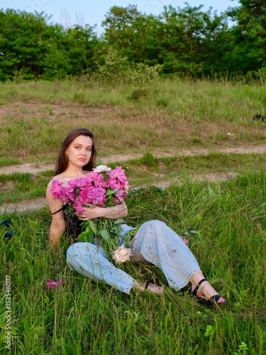 Happy smiling young woman in summer outdoors in a field with pink peony flowers enjoying life in a moment at sunset