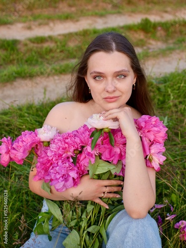 Happy smiling young woman in summer outdoors in a field with pink peony flowers enjoying life in a moment at sunset