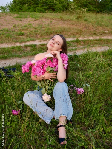 Happy smiling young woman in summer outdoors in a field with pink peony flowers enjoying life in a moment at sunset