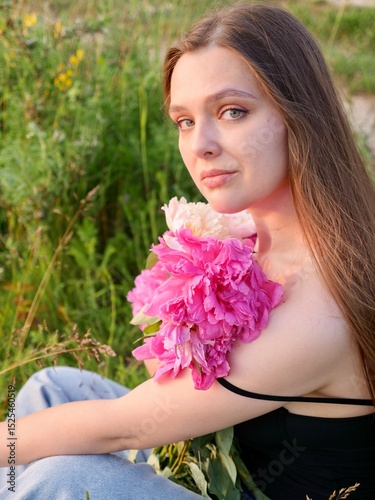 Happy smiling young woman in summer outdoors in a field with pink peony flowers enjoying life in a moment at sunset