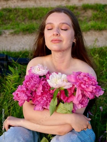Happy smiling young woman in summer outdoors in a field with pink peony flowers enjoying life in a moment at sunset
