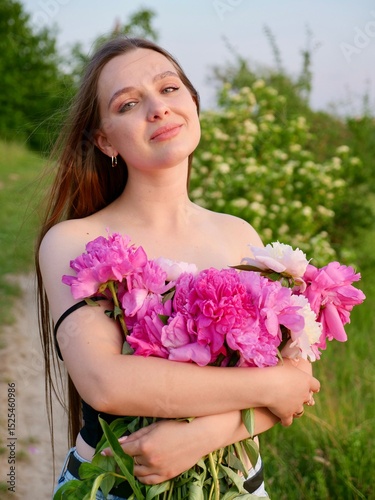 Happy smiling young woman in summer outdoors in a field with pink peony flowers enjoying life in a moment at sunset
