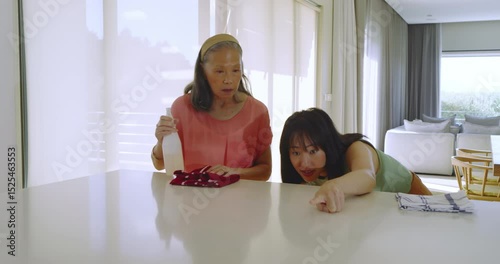 Senior Asian grandmother spraying countertop, granddaughter spotting stain, laughing and cleaning