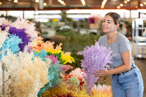 Cheerful young girl choosing branches of preserved Italian ruscus with delicate foliage painted in bright colors at flower market to use in floral arrangement photo