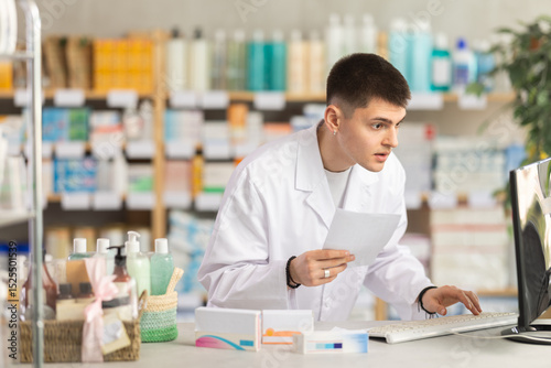 Professional young man pharmacist standing at prescription counter and working on computer, stocktaking medicines at small pharmacy shop