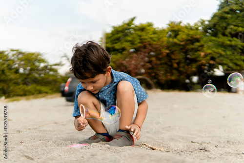 Young boy crouching on sand, making soap bubbles on a sunny day
