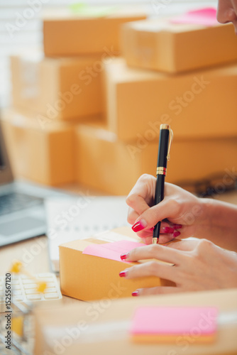 Asian female small business owner working from home taking orders and shipping to customers with laptop, pen and notepad. Ecommerce concept for SMEs.
