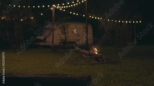 Camper is enjoying a bonfire near his cozy travel trailer in an autumn forest at night, Serene scene of a caravan with a campfire and string lights surrounding, set in a calming forest at dusk