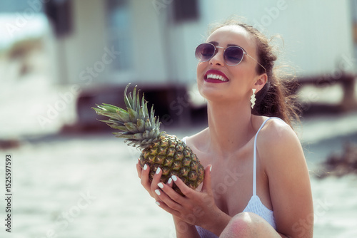 Laughing woman in sunglasses holding pineapple on the beach