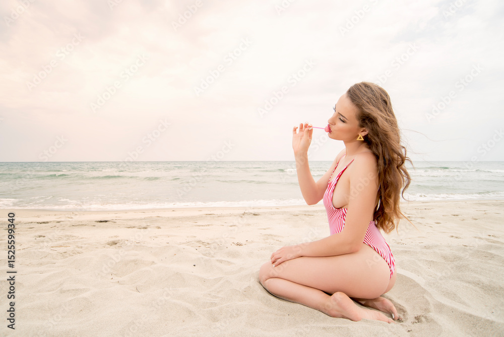 Playful woman in red striped swimsuit kneeling on sand with lollipop Playful woman in red striped swimsuit kneeling on sand with lollipop