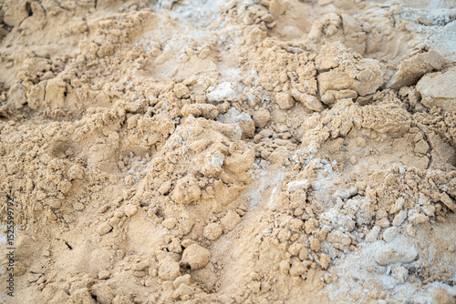 A close-up shot of textured sand, mixed with some small rocks and lighter material, fills the frame. The focus is on the details of the sands surface.
