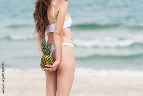 Woman in white bikini holding pineapple behind back on the beach