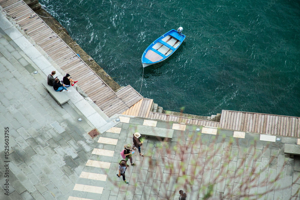 Obraz premium Aerial view of a blue rowboat moored on the Mediterranean coast, with people walking and sitting on a stone promenade. Captures a peaceful seaside moment in an Italian village