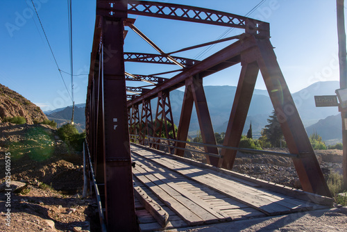 Iron bridge in Tilcara, Jujuy Province, Argentina, spanning a rugged landscape with rust-colored metal beams, set against the backdrop of the Andean mountains. photo