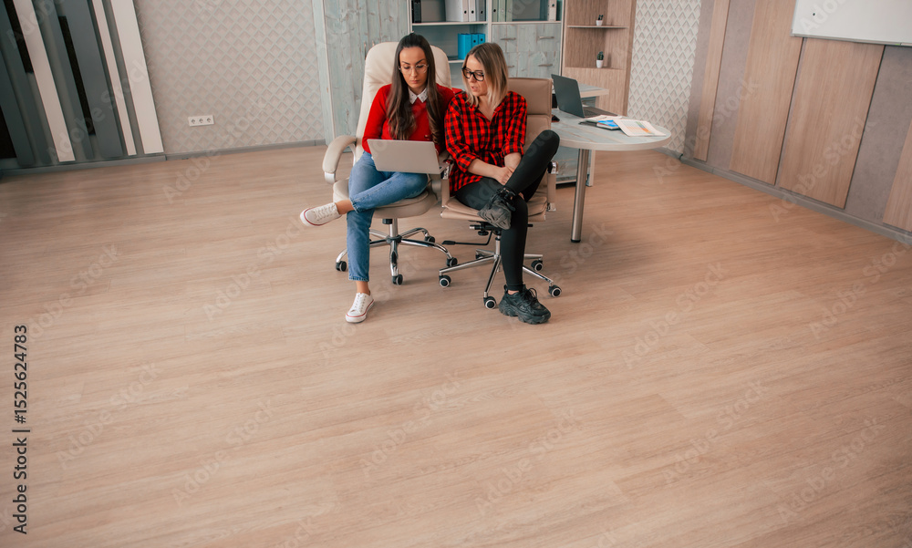 Obraz premium Two young women in casual office wear sitting in swivel chairs, discussing work on a laptop, in a modern office with light wood flooring and minimalist decor.