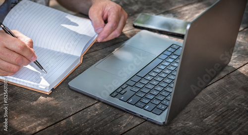 Businessman or freelancer writing in notebook while working outdoors on laptop at wooden table. Creative remote work or planning concept