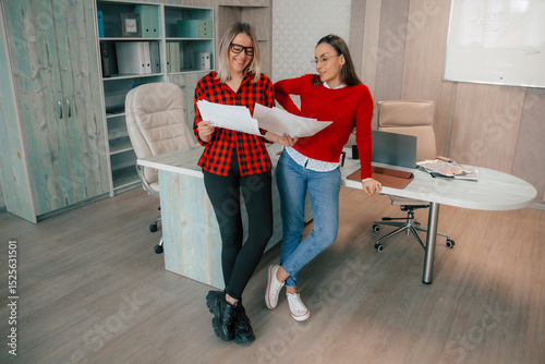 Two young women in casual office wear review documents together, smiling, in a modern workspace with wooden furniture, laptops, and office supplies, showing collaboration and teamwork.. photo