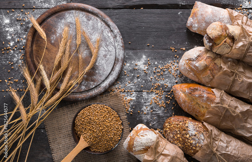 Bread background. Brown and white whole grain loaves wrapped in kraft paper composition on rustic dark wood with empty desk copy space. Baking and home bread making concept. Soft toning