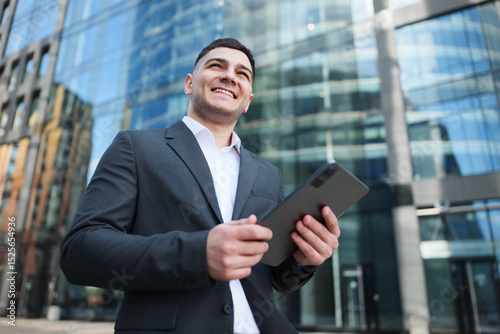 Professional Young Man Smiles While Using a Tablet Outside a Modern Skyscraper on a Sunny Day