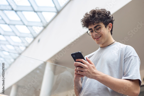 Smiling teenage boy using smartphone in modern urban space. Concept of youth lifestyle, digital communication, mobile technology, and social media engagement. texting, browsing social media, chatting
