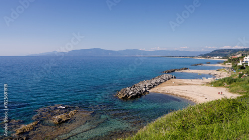 Tranquil Pizzo coastline with crystal clear turquoise waters, featuring a rock barrier and distant mountains on a sunny day