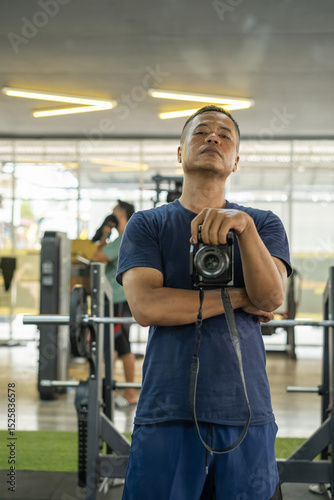 Confident man taking mirror selfie with camera in modern gym. Fitness background with exercise equipment and soft lighting. Urban lifestyle and self-portrait concept in workout space.
