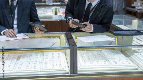 Two jewelry store employees in formal suits checking ring inventory with a clipboard and tablet at a well-lit display counter filled with elegant rings.
