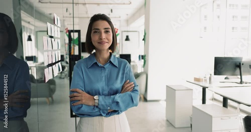Serious confident young Hispanic business professional woman standing at Kanban scrum board in office boardroom, keeping hands folded in confidence gesture, getting happy. Corporate portrait