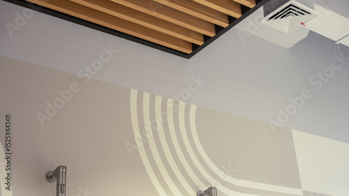 Interior detail of a modern ceiling corner featuring wooden slats, geometric wall pattern, air vent, and minimalist architectural design in neutral tones.