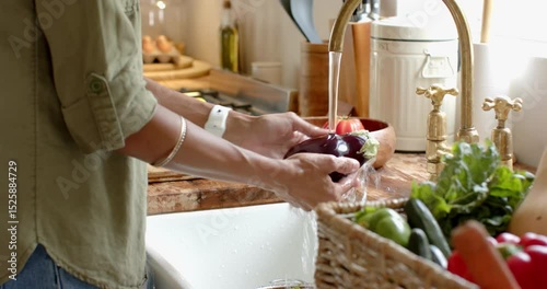 Washing fresh green apple in kitchen sink with vegetables in basket nearby, at home