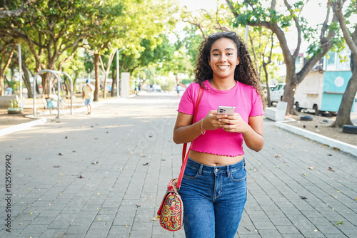 Young latin woman walking in the park and using her smartphone