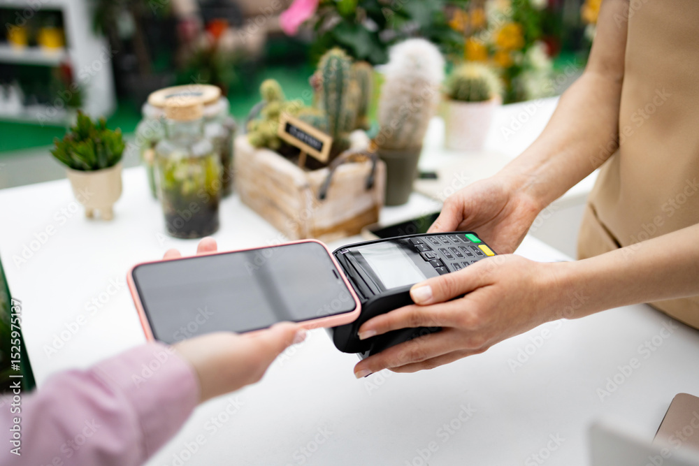 © sofiko14 - A customer pays with a phone using contactless payment at a florist shop. Various plants are visible in the background.
