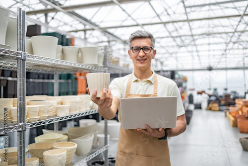 Wallpaper Mural A shopkeeper holding a flowerpot and laptop, surrounded by various ceramic pots, in a plant shop. The man is smiling and wearing an apron. Torontodigital.ca